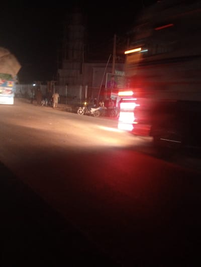 The media file depicts a nighttime street scene in Tundun Wada South, Nigeria, likely during a busy period due to the presence of vehicles and pedestrians. The overall atmosphere is one of motion and activity, suggested by the motion blur evident in many elements of the image, especially the bright red lights on the right side which are likely from a vehicle.
In the foreground, a street is visible, illuminated by artificial lights. The road surface appears to be paved and somewhat worn. To the left, a portion of a large truck or bus is visible, its side showing some colorful decals or advertisements. Further down the street, there are several people. Some are standing, while others appear to be on a motorcycle. The figures are indistinct due to the low light and motion blur, but they convey a sense of everyday life and movement.
In the background, a building structure, possibly under construction or a religious building like a minaret, is visible against the dark night sky. Scaffolding suggests ongoing work. Adjacent to this structure, there's a corrugated metal fence. A signpost with some text is also visible on the right side of the street, though the text is illegible due to the image quality and motion blur.
The scene is illuminated by various light sources: the headlights of vehicles, streetlights, and the bright red lights that dominate the right side of the frame. The time of day is clearly night. The weather appears to be clear, with no indication of rain or adverse conditions. The location cues, such as the architecture and the vehicle types, are consistent with a Nigerian setting, specifically Tundun Wada South. The overall emotion conveyed is one of a dynamic, possibly bustling, urban environment at night, with people going about their business amidst traffic and illuminated structures.
