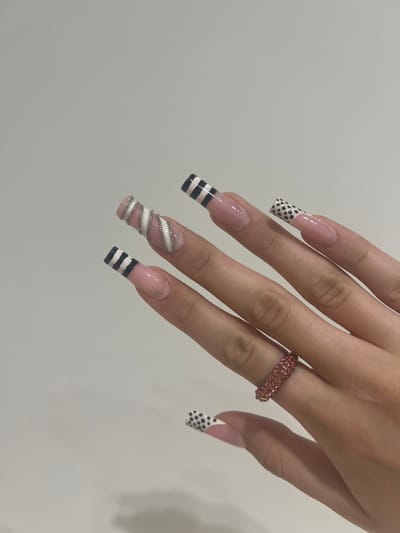 A close-up shot shows a person's left hand with elaborate acrylic nails, against a plain, light-colored background, suggesting an indoor setting, possibly a home or salon, in the city of Искра, Russia. The nails are long and square-shaped, with french tips and intricate designs. The thumb and index finger nails feature a black and white striped pattern. The middle finger nail is adorned with a silver spiral design wrapped around a pink base. The ring finger nail has a black and white polka dot pattern. A sparkling pink ring with multiple small stones is worn on the ring finger. The overall presentation is neat and artistic, highlighting the detailed nail art.