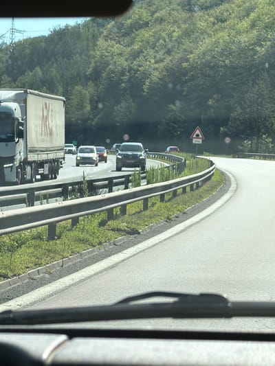The image depicts a highway scene, likely on a sunny day, with dense green foliage covering a hillside on the left and right sides of the road. The road itself appears to be a multi-lane highway with a clear lane division.
In the foreground, the windshield and dashboard of a vehicle are visible, suggesting the photo was taken from inside a car. A guardrail runs along the left side of the road, separating the highway from the grassy embankment.
Several vehicles are present on the highway. A large white semi-truck with a trailer is in the left lane. Further ahead, a black SUV is in the right lane. Other cars, including a white hatchback and a red sedan, are also visible in the distance.
Traffic signs are present. A triangular sign with an image of a tunnel and text "1000m" is visible, indicating an upcoming tunnel. A circular sign with "120" suggests a speed limit.
The environment is natural and mountainous, with a clear blue sky. The presence of a power line tower in the background suggests proximity to infrastructure. No specific landmarks or clues point to the city of Churek, Bulgaria, in this image.