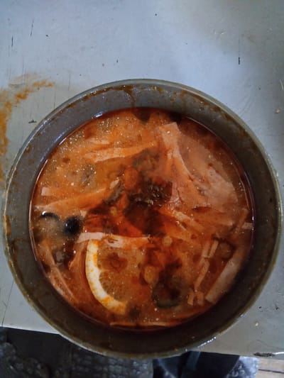 A top-down, medium shot captures a gray, round pot filled with a reddish-orange soup. The soup contains various ingredients, including what appear to be thin strips of meat or vegetables, some leafy herbs, and a slice of lemon with visible seeds. The surface of the soup has a slightly oily sheen with small white specks, possibly spices or fat. The pot is placed on a light gray, textured surface that shows signs of wear and some reddish-orange staining, consistent with spilled food. The background is slightly out of focus but reveals a similar textured surface and what appears to be dark, textured material below the surface. The scene appears to be indoors, in what could be a kitchen or dining area, with no specific indicators of being in Raygorodok, Kazakhstan.