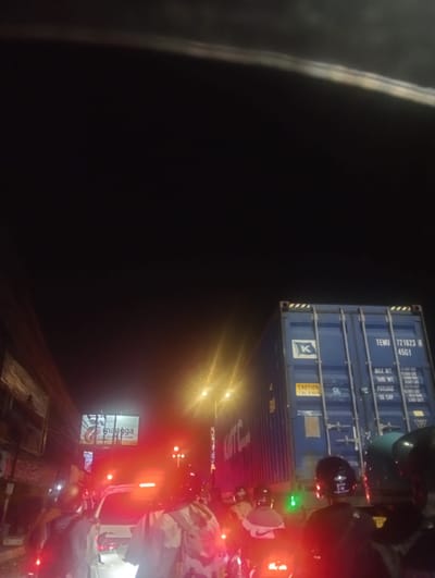 This media file depicts a nighttime scene on a busy street in Calukan RT 1 RW 5 Keboansikep, Indonesia. The primary subjects are people on motorcycles, seen from behind, navigating through traffic. The setting is urban, characterized by road signs, streetlights, and illuminated billboards. A large blue shipping container is a prominent object in the scene, displaying visible text such as "TEMU 721623 4 45G1", "CAUTION 2.9m HIGH", and weight specifications. Another billboard visible in the background has the text "mangga". The overall atmosphere is one of congested urban movement at night, with the red glow of taillights and the bright lights of vehicles and signs illuminating the dark sky. The scene captures the hustle and bustle of a street in Indonesia after dark.