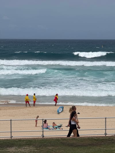 A beach scene shows a wide expanse of ocean with breaking waves meeting the sandy shore. Several lifeguards in red and yellow uniforms are patrolling the shoreline. A group of people is sunbathing and relaxing on the beach. Two women stand near a fence, apparently observing the ocean. The sky is overcast, and a small sailboat is visible in the distance. A grassy area borders the beach behind the fence.