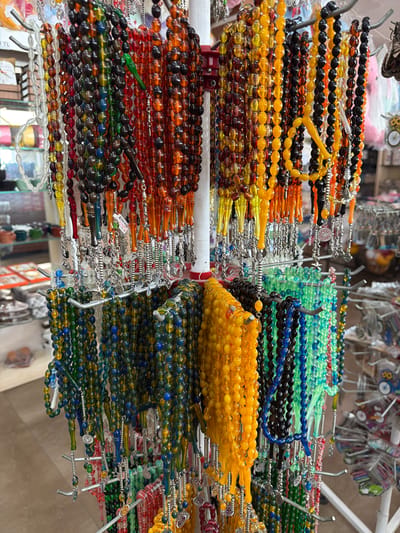 This is a medium-shot, eye-level photograph of a display of prayer beads, also known as tesbih, for sale in a shop. The beads are arranged in various colors and sizes on metal racks. The tesbih are mostly made of smooth, polished stones and glass, in hues of orange, yellow, green, blue, black, and red. Some have silver decorative charms or tassels hanging from them. The background is blurred, but appears to be the interior of a shop with shelves displaying other items. This scene is likely located in the city of Acıpayam, Turkey, a region known for its traditional crafts and souvenirs. There are no people visible in the image.