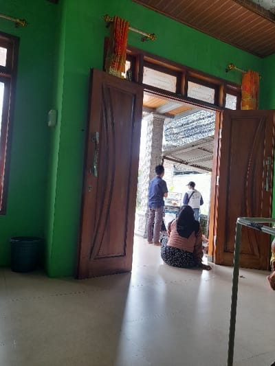 The image depicts an interior scene of a home located in Kecamatan Purwoasri, Indonesia, at coordinates -7.635, 112.136. The room is primarily green with a tiled floor. A large, open doorway leads to an exterior courtyard with a tiled floor. There are three individuals in the courtyard; two standing and one sitting. The standing individuals appear to be males, one wearing a blue shirt and the other a white shirt. The sitting individual is a woman wearing a headscarf and patterned dress. The room contains a brown door on the left side with a window behind it. On the right side, there is another door leading to the courtyard and a chair. A brown, patterned curtain hangs above the doorway on the right, and a black trash can sits in the corner of the room. The light source appears to be natural sunlight coming from the doorway. The interior is sparsely furnished, and the mood is one of normalcy.