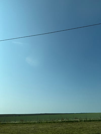 The image displays a vast, clear blue sky with a thin, dark horizontal line, possibly a wire or cable, bisecting the upper portion of the frame. Below the sky, a flat, green landscape stretches to the horizon. This landscape consists of fields of varying shades of green, with some areas appearing as cultivated crops and others as grassy meadows. A faint line of trees is visible on the distant horizon. In the foreground, a strip of mown grass is present, and a wire fence runs across the mid-ground of the image, separating the different fields. The scene is devoid of any people or discernible structures, giving it a natural, open-air feel. The location is identified as Ken'shevo, Russia, suggesting a rural or semi-rural setting.