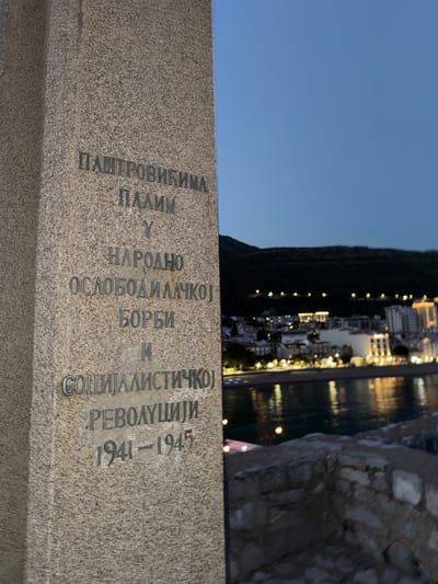 The image depicts a commemorative stone monument in Gornji Katun, Montenegro. The inscription on the monument is in Cyrillic script and reads: "PAŠTROVIĆIMA PALIM U NARODNO OSLOBODILAČKOJ BORBI I SOCIJALISTIČKOJ REVOLUCIJI 1941-1945". In the background, one can see a coastal city illuminated at night, reflecting in the water. To the right of the monument is what seems to be part of a stone structure.