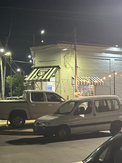 A sideways night shot features a white building with string lights. A black and white striped awning and flag are visible. "CAMPO CHICO" is printed on the flag with a soccer ball logo. Two white vehicles and a black vehicle are parked in front of the building. Streetlights illuminate the scene, and power lines run across the sky.