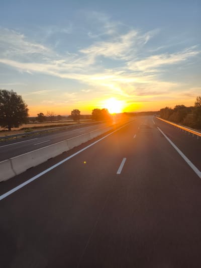 The image shows a highway in Thiel-sur-Acolin, France, at sunset. The sun is low in the sky, casting a warm glow over the landscape and reflecting off the road. The highway is paved and has white lane markings. There are concrete barriers on the side of the road, and a metal guardrail further out. Trees and fields line the highway, with a few scattered trees visible in the distance. The sky is a mix of blue and orange hues, with some wispy clouds. The perspective suggests the photo was taken from a moving vehicle.