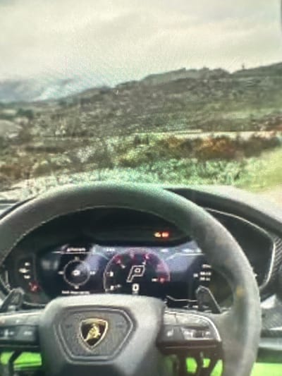 The image shows the interior of a Lamborghini, focusing on the steering wheel, dashboard, and a glimpse of the outside environment. The steering wheel is prominently displayed with a Lamborghini logo on the center. The dashboard is digital and displays various gauges and indicators. The outside view shows a blurred landscape of what appears to be a rural or mountainous area. The location mentioned is Kukorevo, Bulgaria, but that is not verifiable from the image alone.