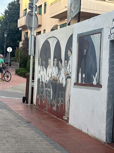 This image captures a street scene in Calella De Palafrugell, Spain, featuring a large mural on a building's white exterior wall.
The mural, centrally located, depicts an interior tavern scene. The left segment shows eight men: seven are seated around a square table, and one stands to the right of the table, appearing to serve. All men wear white shirts and dark trousers; several hold glasses or bottles. The background of this section features dark arched alcoves. The right segment of the mural portrays a painted window-like alcove containing a white wine bottle, a glass with red liquid, and a white, abstract vase or sculpture on a ledge. A dark, hanging lantern is depicted within this window scene.
The immediate foreground consists of a pedestrian sidewalk paved with reddish-brown square tiles directly adjacent to the mural wall, transitioning to grey interlocking pavers further left. A dark brown wheeled trash bin stands near a metal signpost on the reddish-brown tiled section.
To the left, a person wearing a green short-sleeved shirt, dark shorts, and a pink and white helmet is partially visible, walking or standing next to a black bicycle with pink accents on its frame and wheels. Behind them, an asphalt road is visible, flanked by a black street lamp and a white circular road sign with a horizontal bar. Green trees and shrubs line the background, with a multi-story, light-colored residential building featuring balconies and green window shutters visible beyond the foliage.
Above the mural on the right, partial text reading "CA" and "DE" is visible on the building wall. Overhead wires and a billboard with an abstract design are present in the upper background. The scene is illuminated by natural daylight.