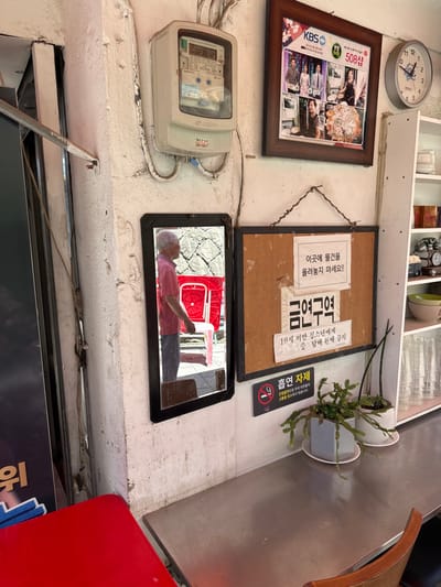 The image captures the interior corner of a commercial establishment, likely a restaurant or bar, located in Jongno-gu, South Korea. A cream-colored electricity meter, displaying "0238.12," is mounted high on a textured, white wall. Below it, a black-framed mirror reflects the profile of an elderly male with white hair, wearing a pink shirt and blue trousers. He is positioned outdoors, with a red plastic chair and a grey, stone-textured wall visible in the reflection.
To the right of the mirror, a large cork board with a dark frame is affixed to the wall, displaying two white notices in Korean. The top notice states, "이곳에 물건을 올려놓지 마세요!!" (Do not place items here!!). The larger bottom notice reads "금연구역" (No Smoking Zone), followed by "19세 미만 청소년에게 술 담배 판매 금지" (Sale of alcohol and tobacco to minors under 19 is prohibited). Below the cork board, a smaller black sign with a crossed-out cigarette icon indicates "흡연 자제" (Please refrain from smoking).
Above the cork board, a dark brown framed picture features multiple smaller images, including individuals and food, with "KBS" and "508쌈" visible. To the right of this frame, a white circular wall clock displays the time as approximately 1:35.
A polished metallic counter, likely stainless steel, extends from the wall to the right, supporting two potted plants. A wooden chair with a curved back is partially visible beneath the counter. In the lower-left foreground, a portion of a red-topped stool or small table is present. A white open shelving unit on the far right holds multiple clear glasses and other items. The wall itself appears worn, with some peeling paint and grime, suggesting age and use.