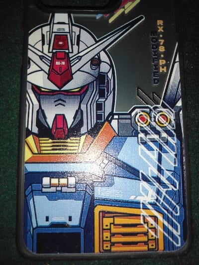 The image displays a close-up of a smartphone case, photographed in the city of Quesada, Costa Rica. The case features a prominent, stylized illustration of the RX-78-2 Gundam's head and upper torso. The design utilizes a color scheme of white, red, blue, yellow, and black outlining. Vertical text on the right side of the illustration reads "RX-78-PH" and "MODIFIED." The case rests on a dark green, textured background, consistent with a fabric surface. No individuals, active subjects, or dynamic interactions are depicted.