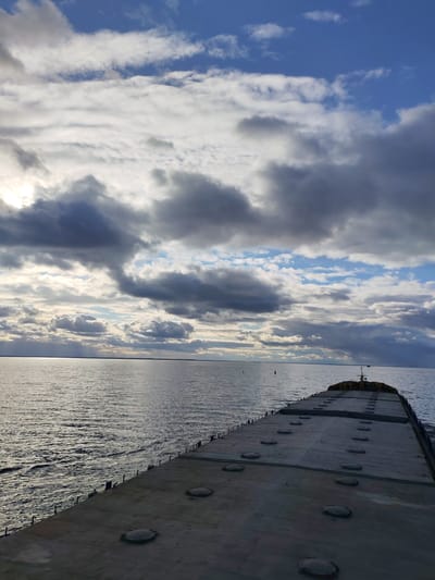 The image displays a wide-angle view from the stern of a large, flat-deck cargo vessel, likely a barge or bulk carrier, traversing a vast body of water. The deck, painted in a dark gray, occupies the lower portion of the frame and features numerous circular hatch covers arranged in rows, receding towards the distant bow. A railing with stanchions is visible along the starboard side of the vessel, where the water churns slightly.

The expansive body of water, which appears to be a large river or reservoir based on the provided location context of Novaya Svobodka, Russia, stretches to a low horizon. The water surface shows gentle ripples and reflects the overcast sky. In the mid-distance on the water, two buoys are visible, indicating a marked waterway.

The sky is dramatic and dynamic, filled with a mix of heavy, textured gray and white clouds that partially obscure the sun. Patches of brighter blue sky are visible in the upper right. On the left horizon, sunlight breaks through the clouds, creating a bright glare on the water and subtle crepuscular rays. The lighting suggests either late afternoon or early morning. No people or visible text are present in the image. The scene evokes a sense of journey and the vastness of the natural environment under a varied sky.