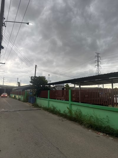 A cloudy day in Na Kluea, Thailand. The image, taken at ground level, depicts a street running alongside a long, green fence with brown wooden slats at the top. Overgrown vegetation grows along the base of the fence. Power lines and poles stretch across the sky. A tall, metal electricity pylon stands near the fence. A streetlight is on the left. A tree is near the fence. A car is visible in the distance. The street is made of asphalt. A blue trash can is by the fence.