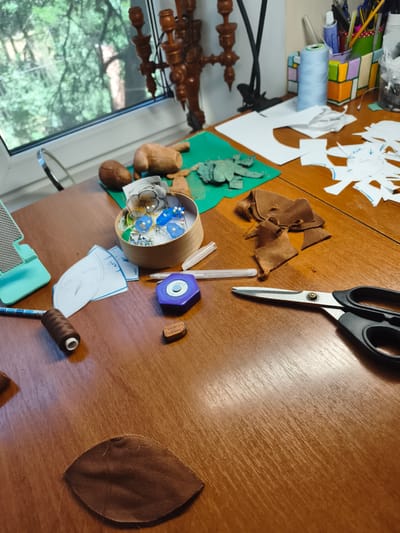 The image depicts a wooden table in what appears to be a craft or sewing area, possibly in Sokolivske, Ukraine. Scattered across the table are various crafting supplies and materials. In the foreground, a pair of black-handled scissors rests with its blades open. To the left of the scissors, a brown spool of thread and a piece of brown fabric cut into a petal shape are visible. Further left, a light blue holder for a device is present. In the midground, a wooden box contains an assortment of pins and small blue bead-like objects, possibly for crafts. Patterns drawn on white paper are spread across the table, along with scraps of green and brown fabric. In the background, a window reveals greenery and trees, suggesting an outdoor view. On the far right, a container holds pens and markers, and a light blue spool of thread stands upright. A wooden candelabra-like object is also visible in the background, near the window. The overall impression is a workspace for textile crafts, such as making dolls or other fabric items. There are no people visible in the image.