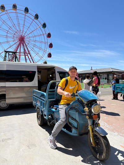 A bright outdoor scene in Jiejinkou, China, captures a man sitting on a blue motorized tricycle, giving a thumbs-up to the camera. He is wearing a yellow t-shirt with a white Nike swoosh, light-colored pants, and a baseball cap. His expression is one of contentment. The tricycle, which appears to be a cargo or utility model, has Chinese characters "精品" (jīngpǐn, meaning "boutique" or "quality product") on its front panel.

In the background, a striking red ferris wheel with multi-colored gondolas (red, green, yellow) dominates the left side of the frame, suggesting an amusement park or fairground setting. To the right, several other people are visible, some standing near a white van or camper, and others closer to some buildings. One person is next to a dark blue cart with Chinese characters "食品" (shípǐn, meaning "food product") on it, indicating a food stall.

The weather is clear and sunny, with a bright blue sky showing only faint, wispy clouds. Strong shadows are cast on the ground, confirming bright daylight. The overall atmosphere appears relaxed and leisurely.