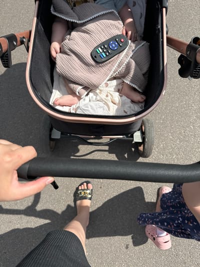 The image depicts a top-down perspective of a person pushing a stroller on a paved surface, likely a sidewalk or road, in Dovhe, Ukraine. The stroller contains a baby, partially covered by a pinkish blanket with white pom-pom trim. A black remote control with colorful buttons rests on the blanket. The stroller has a rose gold frame and black fabric.
The person pushing the stroller is wearing a black skirt and black slide sandals with gold embellishments. Another person, possibly an adult or older child, is walking beside the stroller, wearing a navy blue dress with a colorful pattern and pink crocs. Shadows cast by the individuals and the stroller indicate it is a sunny day. The ground is a typical asphalt or concrete surface found in urban or suburban environments.