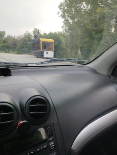 The image shows a view from inside a car, looking out the windshield. The car's dashboard is in the foreground, with air vents and a radio visible. Through the windshield, a road is visible, with a roadside stand selling goods. The stand has a yellow roof and a black body, with items displayed on shelves. Trees and vegetation line the road. The sky is overcast, suggesting a cloudy day. The scene appears to be in a rural or semi-rural area, possibly a marketplace or a place where vendors set up to sell their products. The text "MAXIMA" is visible on the car's radio display. The location is identified as Balta, Uzbekistan. The overall impression is of a casual drive through a scenic area with local commerce.