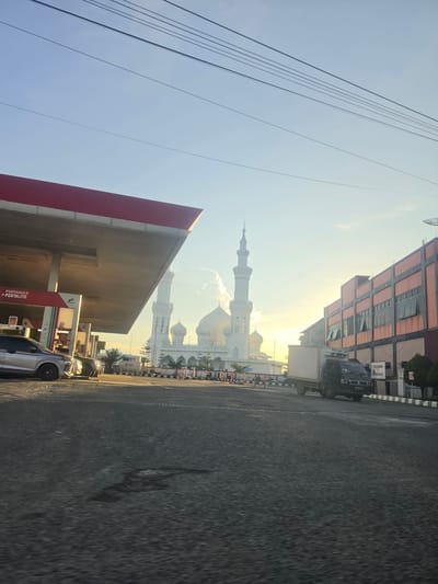 The image, taken in Ranto, Indonesia, presents a street scene with a mosque visible in the background. The foreground includes a gas station with a "PERTAMAX PERTALITE" sign. A silver car is parked at one of the pumps. On the opposite side of the street, there is a multi-story building. Overhead power lines are visible against a clear sky. A truck and several cars are on the road, facing towards the mosque. The overall lighting suggests it is early morning or late afternoon.