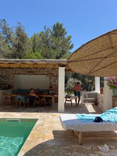 The image depicts a sunny outdoor scene at what appears to be a resort or villa in Santa Eulària des Riu, Spain. A man in green swim trunks is standing near a table, looking towards the camera. He has a towel draped over his shoulder and is barefoot. In the background, several people are seated around a wooden table, engaged in conversation. The setting is rustic, with stone walls, wooden beams, and a large thatched umbrella providing shade. A swimming pool with clear green water is visible in the foreground, reflecting the sunlight. The time of day is daytime, with a clear blue sky, suggesting fair weather. The overall atmosphere seems relaxed and casual, indicative of a vacation or leisure activity. There are no visible text elements in the image.
