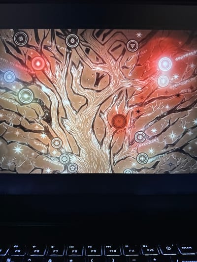 The image shows a close-up of a laptop screen displaying a complex, stylized tree-like diagram. The diagram features interconnected branches with circular icons and glowing orbs placed at various points. The color palette consists of warm tones like orange and red, contrasted with cooler blues and white glows. The background appears to be textured and abstract, possibly representing a mystical or game-related interface. The lower portion of the image shows a dimly lit laptop keyboard. There are no people or recognizable landmarks visible that would indicate a specific location in Vancouver, United States of America.