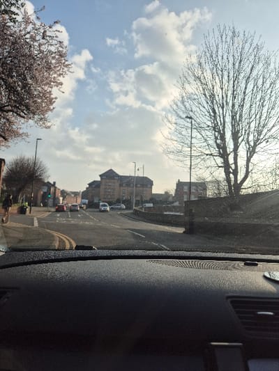 The image depicts a street scene in Hereford, United Kingdom, looking north-east from Newtown Road towards its intersection with Eign Street and Whitecross Road. The prominent multi-story red-brick building in the mid-background, with numerous windows and a gabled roof, is the former Hereford General Hospital, now converted to apartments, located on Eign Street.
The viewpoint is from inside a vehicle, showing a dark, textured dashboard in the immediate foreground, viewed through a windshield exhibiting minor dust and sunlight glare. The asphalt road ahead features a double solid yellow centerline and a dashed white line on the right side. At the intersection, a red hatchback and a dark-colored car are either stopped or turning left. Further along the main road, a white van and another car are visible, proceeding away from the camera.
On the left sidewalk, a pedestrian in dark clothing walks away from the viewer. Above the sidewalk, a cherry tree in bloom displays light pink flowers. A green traffic light is illuminated on a pole to the left of the intersection. On the right side of the road, a low stone wall borders the pavement, behind which a large deciduous tree with bare branches stands silhouetted against the sky. The sky is bright blue with scattered white cumulus clouds. Several dark streetlights are visible throughout the scene.