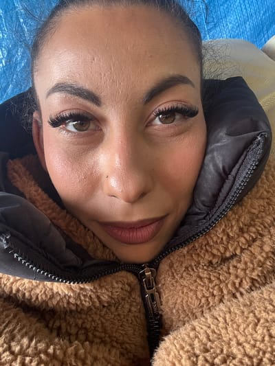 A close-up, slightly rotated, full-face view of a woman. She has long, dark eyelashes, defined eyebrows, and light brown eyes. Her skin has a fair complexion. She wears a dark pink lip color. The woman is positioned close to a brown, sherpa-textured jacket that is partially zipped. The left side of the jacket has a black, puffy material with a zipper running along the edge. A small portion of a blue surface is visible in the background, near the top left corner. The location is not apparent from the image and cannot be determined.