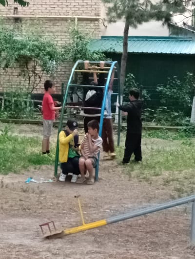 The scene depicts a group of children in an outdoor setting, likely a playground or park in Yukariyuz, Uzbekistan. Five children are visible, interacting around a metal climbing frame. One child wears a red t-shirt and light-colored shorts and stands to the left of the frame. Two other children are seated beneath the frame; one wears a yellow jacket and a baseball cap, and the other is in a striped shirt. Another child is climbing on the frame. A final child, dressed in black, stands slightly apart. In the foreground, a seesaw is visible. The ground appears to be dirt and sparse grass. Behind the climbing frame, there is a brick wall, greenery, and a building with a green roof.