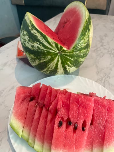 The image displays a close-up view of a watermelon. A large, whole watermelon with a distinctive striped green rind and a slice cut out is positioned in the upper portion of the frame. The exposed flesh of the watermelon is a vibrant pink color. In the foreground, a white plate holds several slices of watermelon, neatly arranged. The slices show the pink flesh and green rind. The scene is set on a marble-patterned surface, likely a table. The background is blurred, showing hints of a teal-colored object and a lighter-colored wall. There is no indication of the city of Bujum, Kyrgyzstan, or any people in the image.