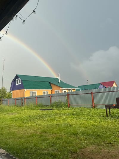 This is a vertical, eye-level, outdoor shot of a rural scene in Южное Снт, Russia. The weather appears to be overcast with a faint rainbow arching across the sky from left to right. The foreground is dominated by a lush, green grassy area, partially mown, with some patches of bare earth. A corrugated metal fence with red posts runs along the right side of the frame, separating the grassy area from the buildings.
Several houses with colorful, corrugated metal roofs are visible. The largest house in the center has a bright orange lower section and a bright green roof, with visible chimneys. A smaller section of this house is painted purple with a single white-framed window. Further back, another house features a red roof. The scene suggests a residential area in a dacha (summer house) community.
In the upper left corner, a string of incandescent light bulbs hangs from what appears to be a wooden structure, adding a decorative touch to the scene. There is no visible text in the image. The overall impression is one of a post-rain, slightly gloomy but picturesque day in the Russian countryside.