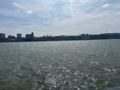 The image depicts a panoramic view of an urban waterfront scene in Cuza Vodă, Romania. The foreground is dominated by a wide body of choppy water, likely a river, reflecting direct sunlight in numerous shimmering patches across its dark, grey-blue surface. No vessels or individuals are visible on the water. A distant shoreline marks the midground, featuring a narrow strip of land sparsely lined with bare deciduous trees and a few small, indistinct structures. The background reveals a low-rise cityscape. On the left, several multi-story concrete buildings, possibly residential or industrial, are present. Towards the center-left, a prominent, multi-story industrial building with a tall, rectangular, silo-like extension on its right side stands out. Further to the right, the skyline continues with more urban structures interspersed among trees. On the far right, a slender, lattice-structured communication tower ascends from a distant, tree-covered hill. The sky overhead is partially cloudy with visible patches of blue, and a strong sun glare emanates from the upper-right quadrant, extending diagonally towards the water.