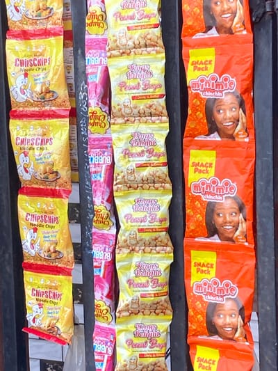 The image presents a close-up view of multiple rows of snack packages displayed on a retail shelf or stall, likely in Sasaro, Nigeria.
The top row features several orange "minimie chinchin SNACK Pack" bags, each adorned with a smiling woman's face and showing pieces of chin chin.
Below this, the second row consists of yellow "Kings Delight Peanut Burger" packages, featuring a cartoon peanut character and marketing text like "Crunchy & Nutritious."
The third row displays pink and yellow "Chupa Chups BUBBLE GUM" lollipops, with "STRAWBERRY" flavor visible on some.
The bottom row showcases yellow "CHIPSCHIPS Noodle Chips" bags, which feature a cartoon chicken and text such as "Chicken Flavor" and "Ready to Eat Crunchy."
The bright, colorful packaging indicates items intended for sale. No specific activity is taking place other than the display of products. Some handwritten text, possibly numbers and letters like "15800010033" or pricing, is visible on a dark support beam between the rows, though it is partially obscured. The scene captures a typical assortment of snacks available in a local market or shop.