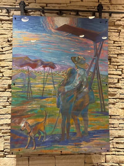 A large, rectangular painting hangs on a wall constructed of stacked, rough-cut light-colored stone. The painting depicts a couple embracing and a dog on a leash in a field with mountainous terrain and a town in the background under a vibrant, multicolored sky. The couple, one person wearing a wide-brimmed hat, stands towards the right side of the painting, their forms rendered in broad, impressionistic strokes of blue, green, and brown. The dog, a slender, tan-colored animal, is positioned in the lower left of the painting, facing left and straining against its leash. In the middle ground, several structures with triangular tops supported by thin poles are visible, casting long shadows on the green and yellow field. Beyond the field, rolling hills and a cluster of buildings suggest a town or city. The sky is a riot of color, with streaks of blue, pink, orange, and yellow, and a large, circular form, possibly the sun or moon, is depicted in the upper right. The painting is illuminated by three circular lights embedded in a dark overhang above it. The stone wall is lit from the side, creating shadows that highlight the texture of the stones. The overall impression is of a serene, artistic depiction of a landscape and its inhabitants.