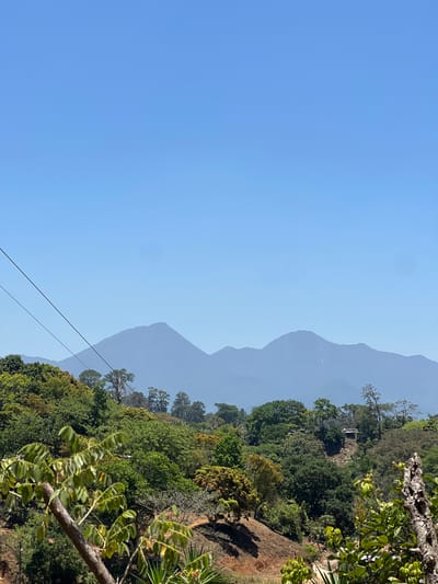 The image shows a landscape in Santa Cruz de Yojoa, Honduras, with mountains in the background. The sky is clear and blue. Lush green vegetation covers the slopes and foreground, including trees and plants. A power line stretches diagonally across the scene. The mountains fade into a hazy blue due to the distance.