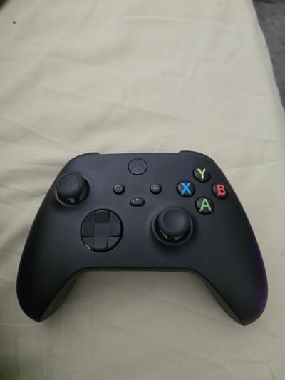The image shows a black Xbox controller placed on a beige, wrinkled fabric surface. The controller is oriented horizontally, with the joysticks and buttons clearly visible. The directional pad is on the left, and the face buttons (A, B, X, Y) are on the right, colored red, green, blue, and yellow, respectively. There are also smaller buttons located between the joysticks and the directional pad. The lighting appears to be overhead, casting soft shadows. No people are present in the image. The background is entirely composed of the fabric surface, which has a slightly textured appearance due to the wrinkles. There is no indication of the city of Worms, Germany, or any specific location within it.