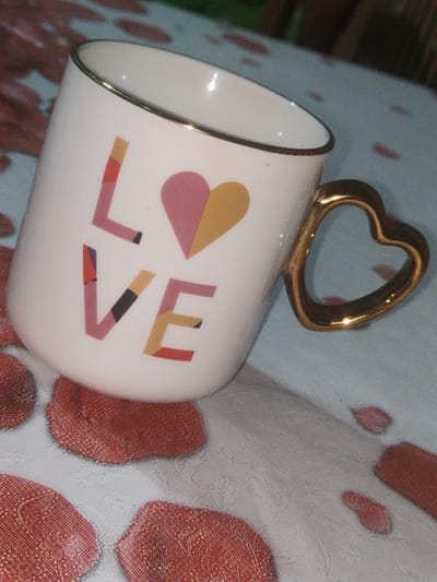 A white mug with a gold heart-shaped handle is shown on a patterned surface. The mug has the word "LOVE" printed on its side in colorful block letters and a heart shape. The surface appears to be a tablecloth with a floral or abstract pattern in red and white. There are also red petal-like shapes scattered on the surface. The scene is lit from above, casting a shadow. The image appears to be taken indoors, possibly in a home. There are no people or animals visible in the image.