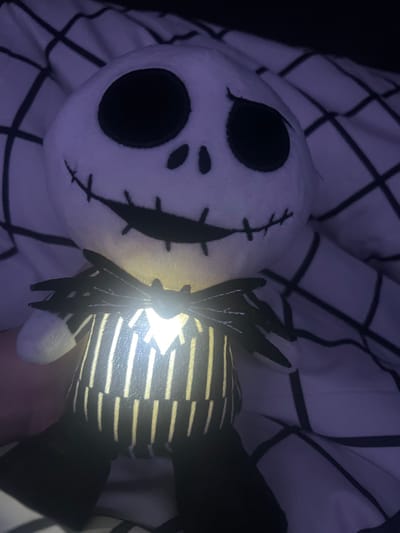 A plush toy resembling Jack Skellington lies against a checkered black and white pattern. The toy's head is white with stitched details forming a skeletal face, including large, black, round eye sockets and a stitched smile. The body has alternating black and white stripes, illuminated from within by a bright, internal light source, visible between its limbs. The scene is dimly lit, giving the image a soft, eerie glow.