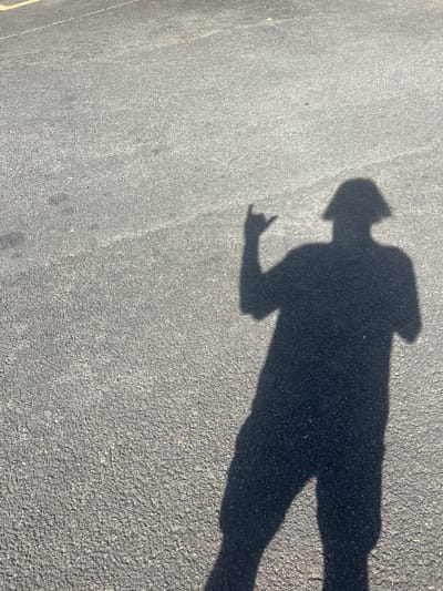 A shadow of a person on an asphalt surface is seen. The person's shadow shows them wearing a hat or hood, with one arm raised in a gesture resembling the "rock on" or "devil horns" hand sign. The surface appears to be pavement, potentially a road or parking lot. The image doesn't contain any direct information linking it to Bellambi, Australia, so any claim of the photo's location is unsupported.