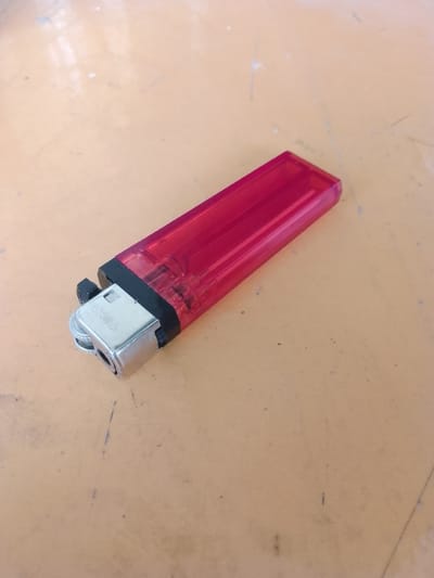A single red plastic lighter rests on a light brown, textured surface. The lighter is oriented diagonally from the bottom left to the top right of the image. The metal spark wheel and igniter are visible on the left side of the lighter, with the word "TOKAI" embossed on the metal. The surface appears to be a table or countertop with some minor scuff marks and a few small dark specks. There are no people or other objects in the frame. The lighting suggests an indoor environment with overhead light sources causing some reflections on the surface. The background is out of focus, but the overall impression is of a simple, everyday setting. This scene could be located in Jonggol, Indonesia, as part of an interior or exterior environment.