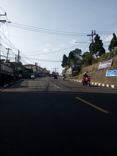 The image shows a street scene in what appears to be a city in Indonesia. Several people are riding motorcycles on a paved road with yellow lane markings. Buildings and power lines are visible on the left side of the road, while a stone wall with banners and trees lines the right side. The banners appear to have holiday greetings written in Indonesian. The sky is light blue with scattered clouds.