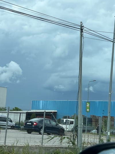 This media file shows an outdoor scene with a blue industrial building in the background. Several utility poles and wires are prominent in the foreground, with a power line stretching across the image. A black car is parked behind a metal fence, and there are stacks of wooden pallets and other industrial materials visible. The sky is cloudy, suggesting overcast weather. A green sign with text in a yellow border is attached to a pole near the building. The image appears to be taken during the daytime. The scene is located in Sânnicoară, Romania, as indicated by the provided location context.