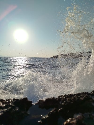 The image captures a dynamic ocean scene near Karaburun, Turkey, under a bright, sunlit sky. In the foreground, dark, wet rocks form a textured boundary. The midground is dominated by the shimmering, dark blue sea, with sunlight glinting off its surface. A powerful wave is captured mid-splash, with a significant volume of white water erupting upwards and outwards, creating a spray of droplets that extend towards the sky. The horizon shows a distant landmass, possibly a coastline or island, silhouetted against the bright sky. The sun, a bright white orb, is visible in the upper left quadrant, casting a strong glare and lens flare that extends downwards and to the left. The sky is a clear, pale blue, with some smaller water droplets suspended in the air, illuminated by the sun. The overall impression is one of natural energy and the raw beauty of the sea.