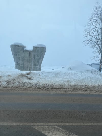 The image presents a stark, overcast winter scene in Borisovo, Russia. A wet, dark asphalt road with a faded white lane marker runs across the foreground, showing signs of dirt and moisture. Adjacent to the road, a substantial bank of dirty, slushy snow is piled. In the midground, to the left, stands a large, unornamented concrete structure, possibly a bridge support or pier, its upper portion featuring a wide, U-shaped indentation and capped with fresh snow. Vertical discolorations and streaks are visible on its concrete surface. To the right, beyond more snow accumulation, a section of a bare deciduous tree rises, with hints of a fence and a building featuring a blue facade visible in the far midground. The entire background is dominated by a uniform, light grey overcast sky. No individuals, vehicles, or specific actions are depicted.