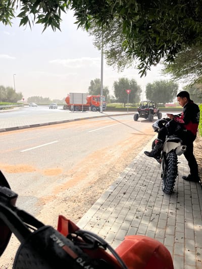 The scene is set in Al Madam, United Arab Emirates, during daylight under a hazy sky. The photograph is taken from the perspective of an all-terrain vehicle, whose red front fairing and black handlebar pad, marked with "RACE," are visible in the lower foreground.

On the right, a male subject stands on a grey paved sidewalk next to a white dirt bike. He wears a black and red long-sleeved riding shirt, black pants, and black boots. His left foot is on the dirt bike's footpeg, and he is observed looking down at a small white object held in both hands. Behind him, off the asphalt road, a black Utility Terrain Vehicle (UTV) with a roll cage is parked on a sandy shoulder. A tall, light-colored utility pole stands to the left of the UTV.

The asphalt road, partially covered with reddish-brown sand patches, occupies the center-left. Two light-colored passenger vehicles are in motion on the road in the distance. Further left, an orange tanker truck, featuring a large tank and a silver, rectangular rear component, is parked on the roadside. The environment includes sandy ground, green trees, and shrubs bordering the road, with a "Give Way" sign visible in the mid-ground. The general illumination is bright, with a soft, dusty light.