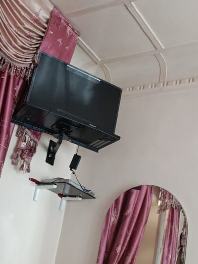 The scene depicts an interior room located in Khasanbay, Uzbekistan. A black Sony flat-screen television is mounted on a light beige wall using a black articulated wall bracket, angled slightly downward. Below the television, a small wall-mounted glass shelf with white brackets and red accents supports a black rectangular electronic device and associated cables. To the left of the television, a section of heavy drapes is visible, featuring an upper light-colored, pleated fabric and a lower maroon fabric with an abstract pattern and decorative tassels. The white ceiling incorporates recessed rectangular panels and decorative molding with dentil-like elements and curved sections. In the lower right of the frame, an unframed arched mirror reflects a portion of the maroon drapes and an indistinct, partial human figure.