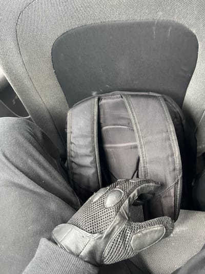 A person is seated inside a vehicle in Pobrdje, Montenegro. The individual is wearing black trousers and a dark long-sleeved garment with a ribbed cuff on the right wrist. A black tactical-style glove, featuring leather palms, mesh backing, and reinforced knuckles, is worn on the right hand. This gloved hand is resting on a black backpack positioned upright between the individual's legs and the back of a gray fabric vehicle seat. The vehicle interior includes textured gray fabric upholstery and a darker, smoother fabric panel visible behind the backpack.