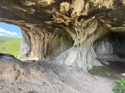 The image depicts a large, natural rock shelter or cave in Skravena, Bulgaria. The cave has a prominent opening on the left side, framing a view of a green, forested hillside and a patch of blue sky with some clouds. The interior of the cave is a complex arrangement of rock formations, with varying textures and colors, ranging from light grey to dark brown and black. The cave floor is uneven, consisting of rock and some patches of grass. The interior is relatively dark, contrasting with the bright exterior view.