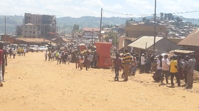 A crowded dirt road scene. People gather and walk along the thoroughfare lined with buildings, some under construction. Cars and trucks are parked along the roadside. In the background, rolling hills and a sprawling town are visible under a bright, sunny sky. A large red structure stands out amidst the crowd. The overall impression is of a bustling marketplace or event in a developing area.
