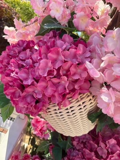 A close-up, vertical outdoor shot shows a woven basket overflowing with artificial pink and fuchsia hydrangea flowers. The basket is positioned on a white wooden structure, possibly a fence or railing, with hints of greenery and foliage in the background. The lighting suggests it is daytime, with sunlight casting shadows and highlighting the delicate texture of the flower petals. The overall impression is one of vibrant floral decoration, likely part of a garden or outdoor display. The scene is devoid of people.