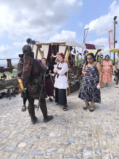 The image depicts a street scene at a medieval fair, likely in Óbidos, Portugal, with cobblestone paving and tents in the background. In the foreground, a person dressed in medieval attire, including a leather tunic, leggings, and boots, is standing with a quiver of arrows on their back. To their right, three women are present. One woman, also in medieval-inspired clothing with a white blouse and dark skirt, is looking towards the archer. Another woman in a black and white floral dress is holding a smartphone and looking down. A third woman in an orange patterned dress is walking away from the camera. Several stalls and displays with medieval-themed items are visible behind the individuals. The sky is partly cloudy, suggesting an outdoor event.